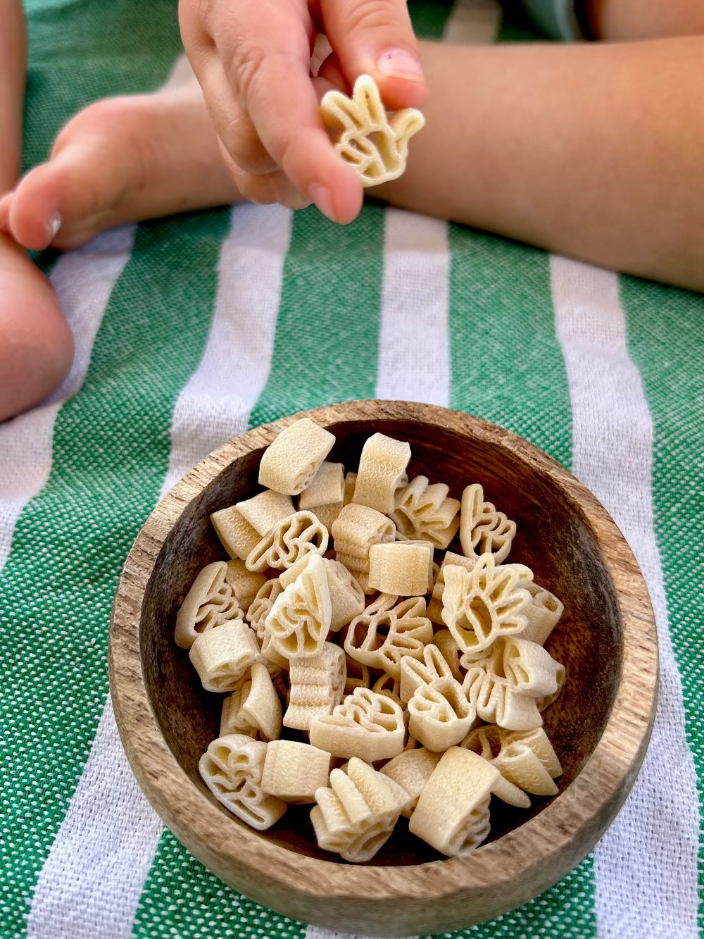 Wooden bowl of 16 oz 1-3-5 American Sign Language Handshaped Pasta. Child holding a pasta piece on green striped cloth